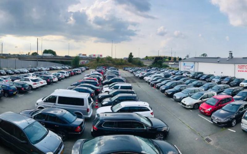 Reihen geparkter Fahrzeuge auf dem Gelände, im Hintergrund Autobahnbrücke und Gewerbebauten